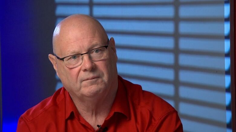 A man wearing glasses and a red shirt sits in front of a blue wall. 