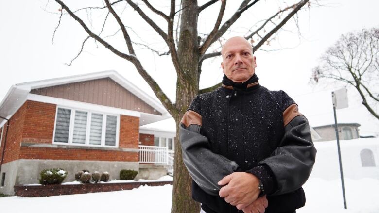 Man, looking serious, stands in front of a tree in a winter scene.