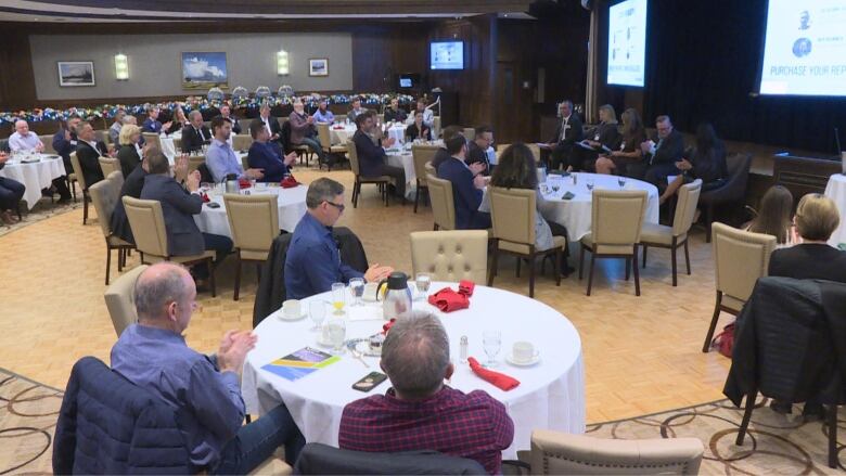 a room of people sitting at tables with white table cloths listening to five people sitting at the front of the room