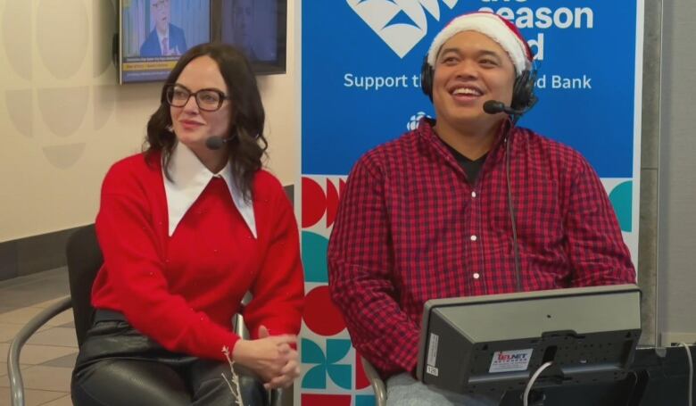 A woman in a red sweater and a man in a plaid shirt sit in chairs and smile. 