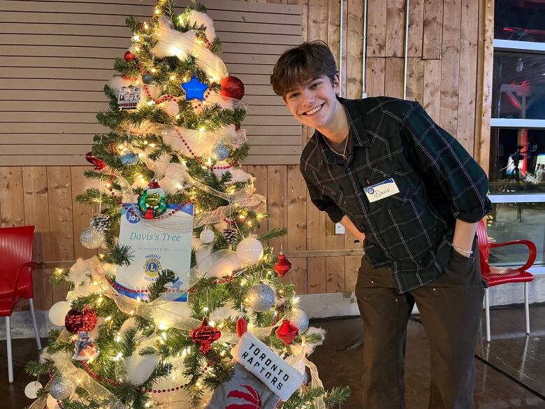 A young boy stands next to a Christmas tree smiling. 