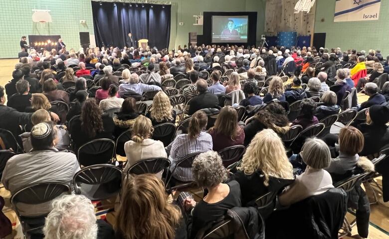 A large crowd of people sit in a gymnasium, looking at a person speaking on a screen.