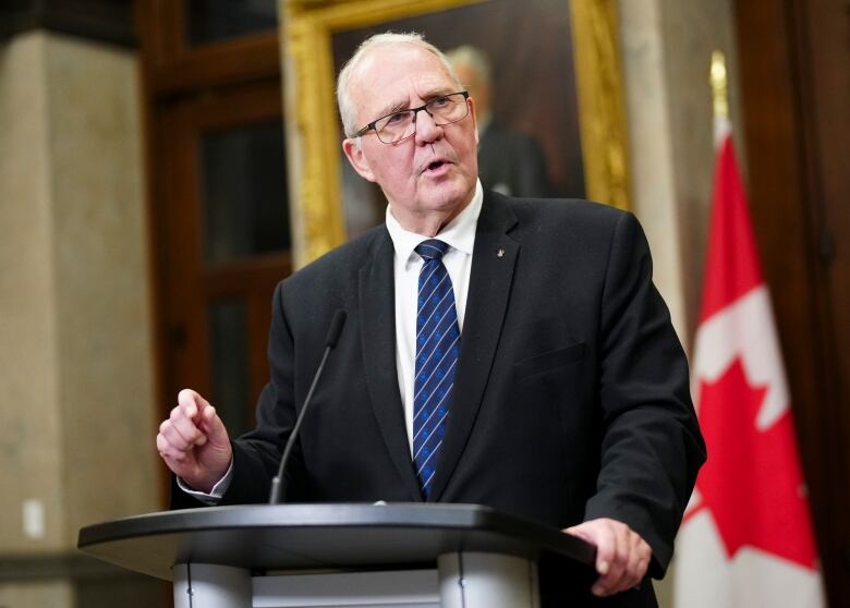 An older man wearing glasses stands at a podium with a Canadian flag behind him.