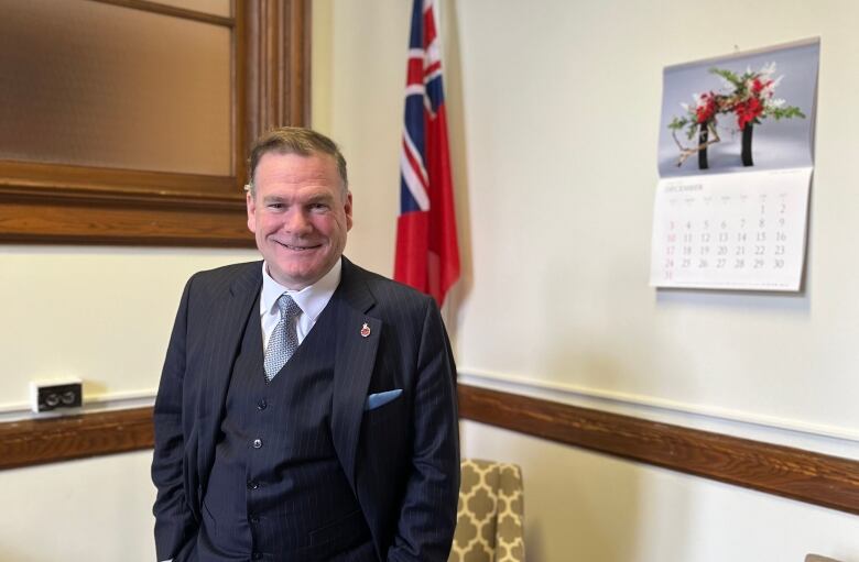 A man in a suit stands in front of a flag.