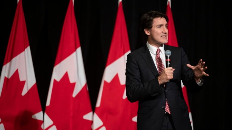 Justin Trudeau wears a black suit and speaks into a microphone in front of a row of Canadian flags.