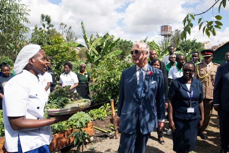 A person speaks to another person holding a shallow basket of green vegetables as they walk through a garden.