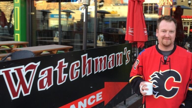 A man in a red Calgary Flames hockey jersey, holding a white mug, stands beside a patio with a sign for the Watchman's pub.