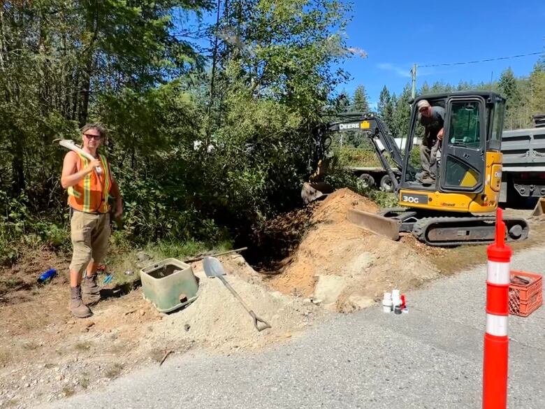 A man on the left in a high visibility vest is holding a wrench over his shoulder and another man on the right is getting out of an excavator. Between them is a hole in the ground to fix pipes. 