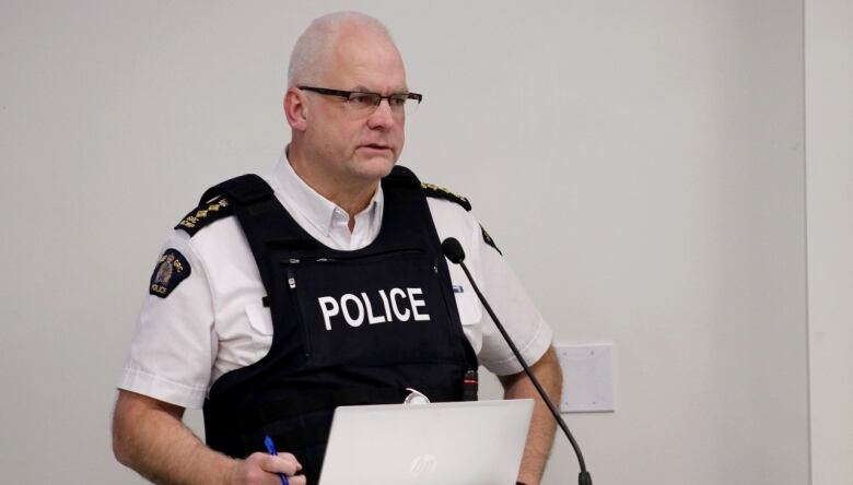 A white man with white hair and glasses stands at a podium in a white shirt and black tactical vest with the word 'police' on the chest