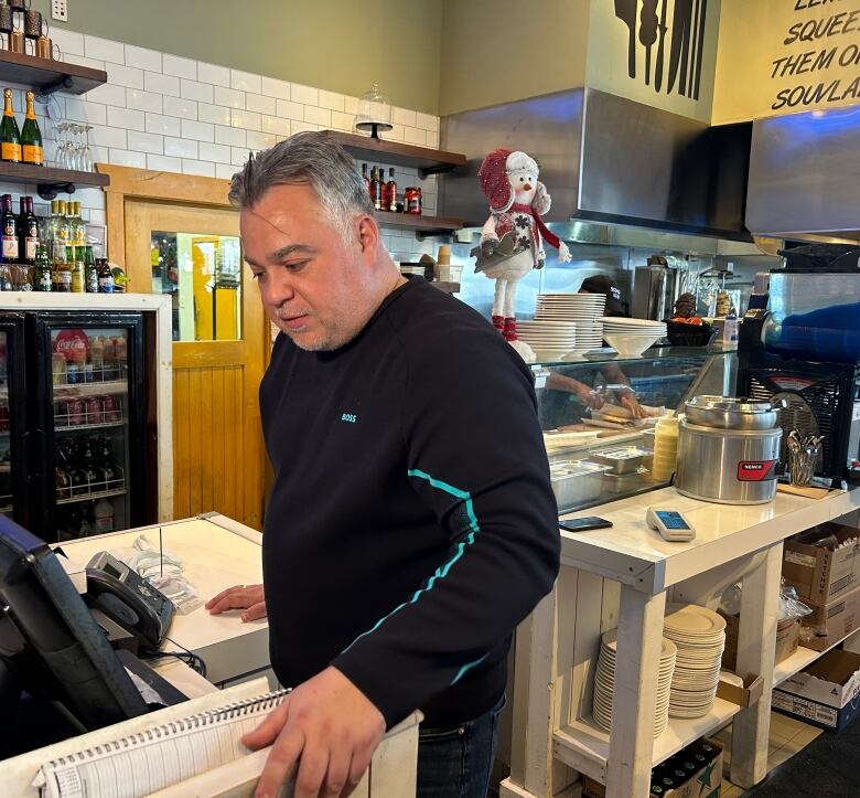 A man stands at a cash register in front of the kitchen of a restaurant.