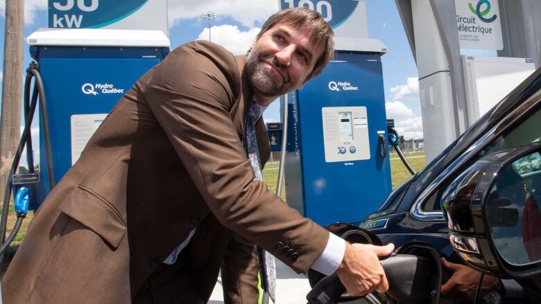 A man wearing a brown suit smiles as he plugs in an electric vehicle at a charging station. 