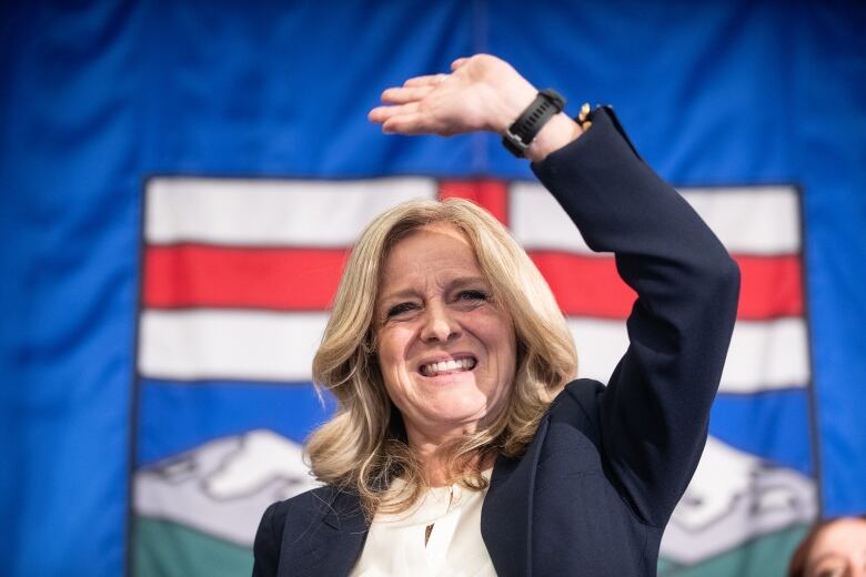 A woman waves over her head. She's standing in front of a giant Alberta flag.