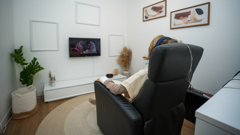 A woman sits in an armchair watching a film with earphones on, as her head is connected to a set of cables behind her.