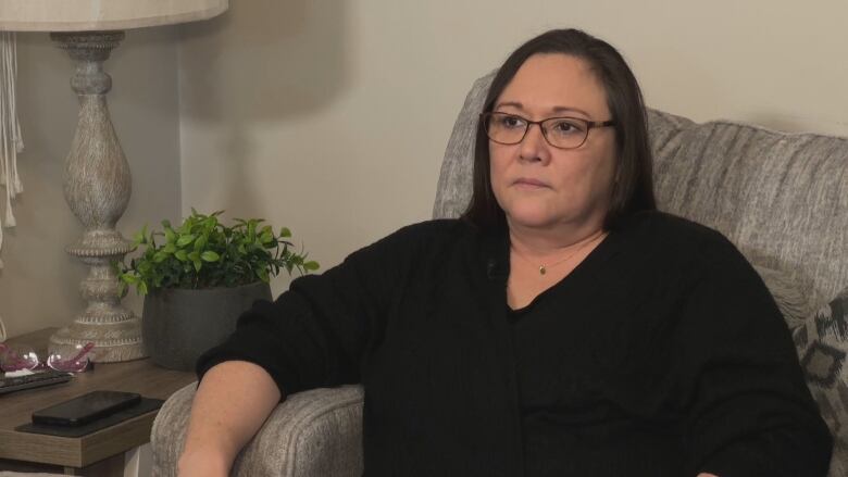 A light-skinned woman with straight dark hair and glasses is wearing a black cardigan and a thin necklace. She is sitting on a couch. There is a houseplant and a lamp sitting on a table to her right.