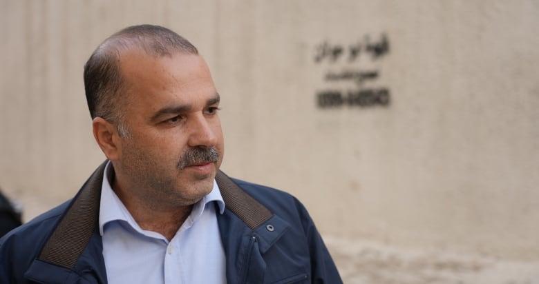 A man with short black hair and a moustache, wearing a navy jacket and light-blue shirt, stands outside a building.