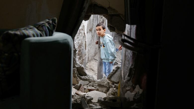 A young boy peers through a large hole in a concrete wall.