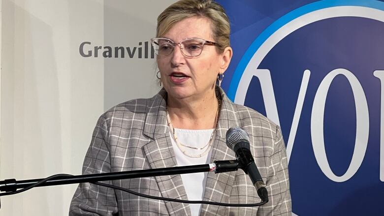 A white woman with blonde hair is wearing glasses and a grey checkered blazer over a white blouse. She's standing at a podium.