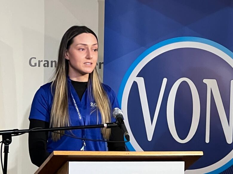 A white woman with blonde and brown hair is standing at a podium in front of a sign that says VON. She's wearing a blue short sleeved t shirt 