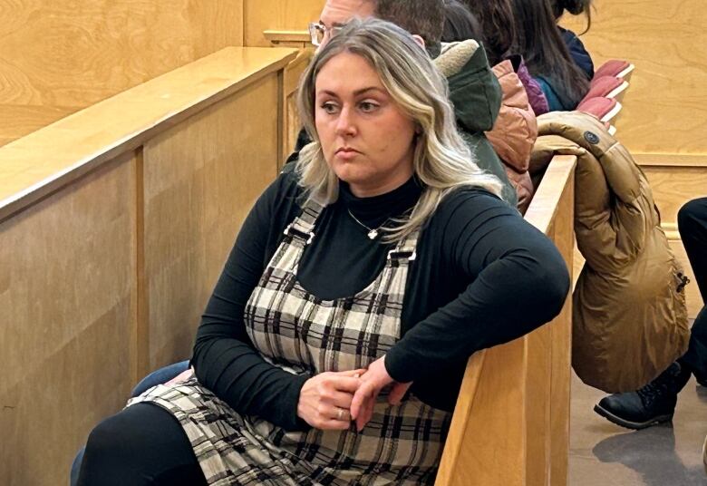 A woman wearing a dress with a black turtleneck shirt sits on a bench in a courtroom.