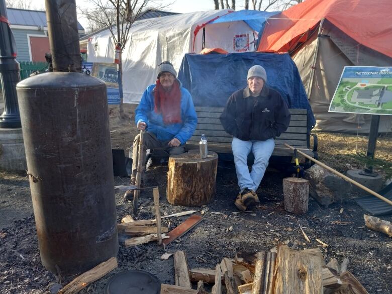 Two men sitting on a bench with a wood furnace boiler set up. Behind them are tents.