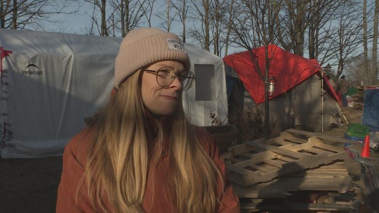 Woman with blond hair, standing outside. Behind her are tents