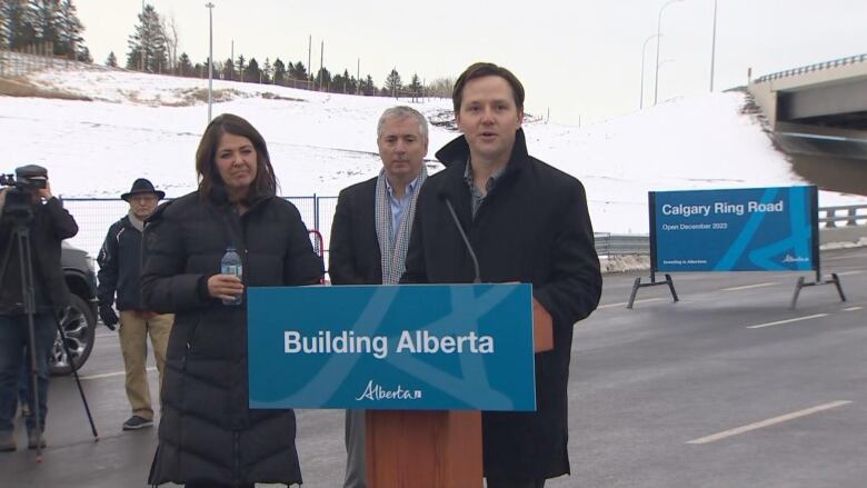 a man in a blue suit and coat stands at a podium with a woman standing beside him and a man behind him on a blocked off roadway