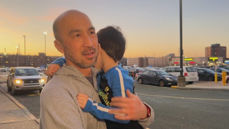 A man with a short beard and mustache is holding a child while standing on a sidewalk. A parking lot is behind them.