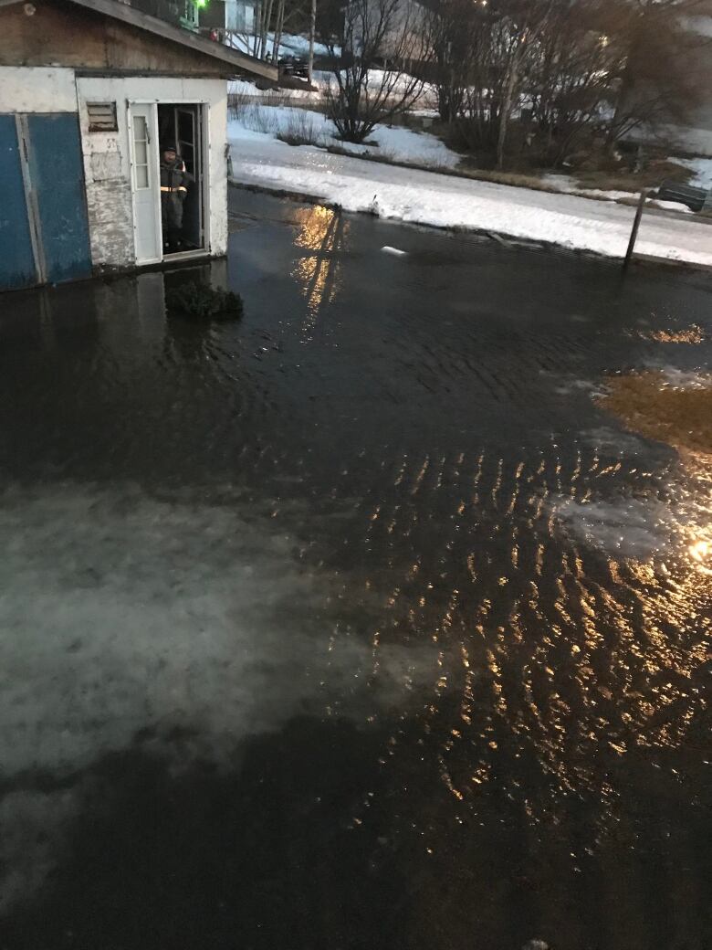 A small shed is surrounded by dark, murky water. A road is in the background. 