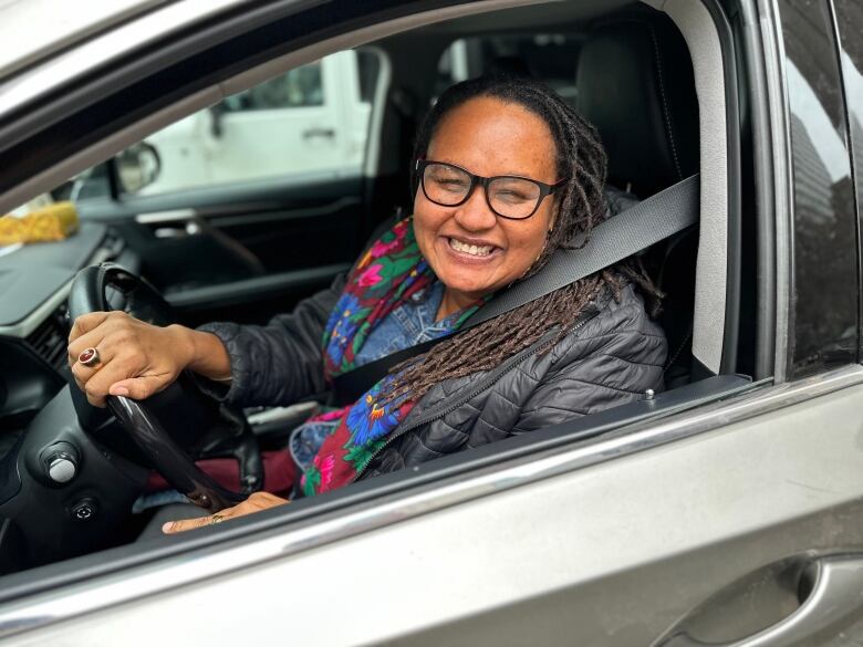 Someone smiles behind the wheel of a grey car.