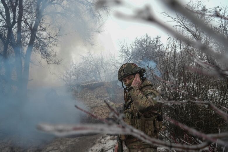 A soldier, framed by tree branches and smoke, covers his left ear with his hand.