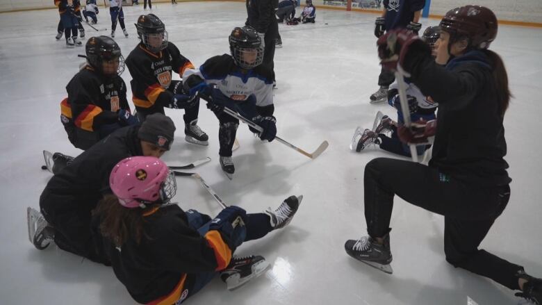 A hockey instructor wearing a helmet and holding a hockey stick is on one knee on the ice. In a circle, there are kids around her wearing hockey gear. 