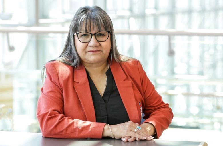 Posed photo of woman with black-and-grey streaked hair, hanging past shoulder length, wearing glasses and a light red jacket over a black blouse.