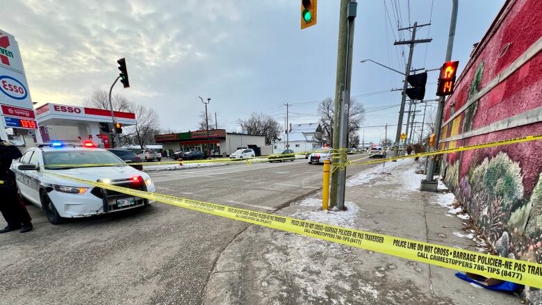 A road is blocked with yellow police tape. A police car is parked in the middle of the road. An Esso gas station and 7-Eleven are in the background.