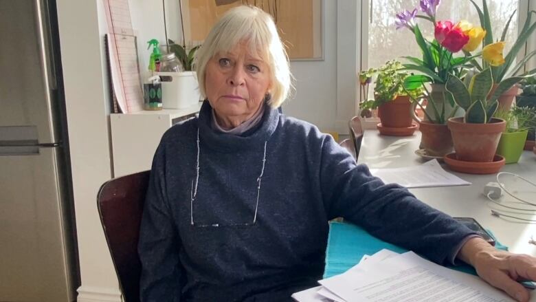 A white woman with a white-blonde bob and vacy sweater sits at a table in a sunny kitchen