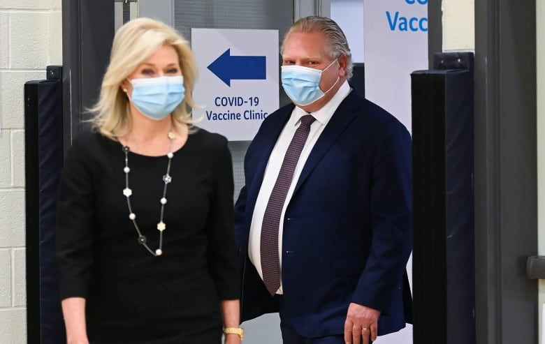 Bonnie Crombie and Doug Ford each wear a surgical mask standing on either side of a sign that says 'COVID-19 Vaccine Clinic.' 