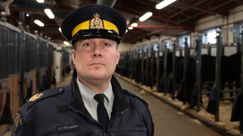 A uniformed police officer with a yellow band on his cap stands in a horse stable.