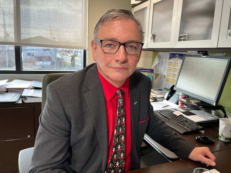 Man in grey suit and red shirt, sitting at a desk.