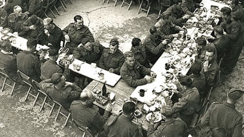 A black and white photo shows soldiers from the Seaforth Highlanders eating their Christmas dinner in 1943 in what looks like a courtyard.