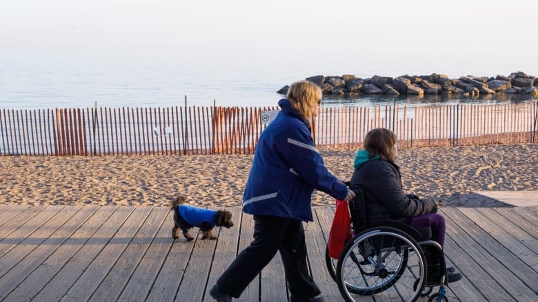 A person pushes another person on a wheelchair, walking close to a body of water.