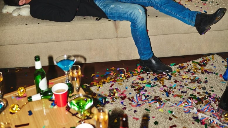 a stock image pictures a person on a couch next to a table full of alcoholic beverages. there is confetti all over the floor and the room is messy.