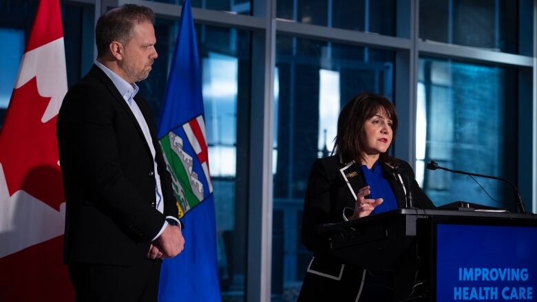 a man and a woman stand at a podium with Canadian and Alberta flags behind them.