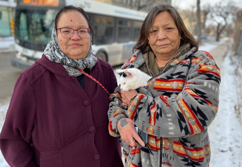 Two women stand outside in the snow. The one at right holds a white cat.