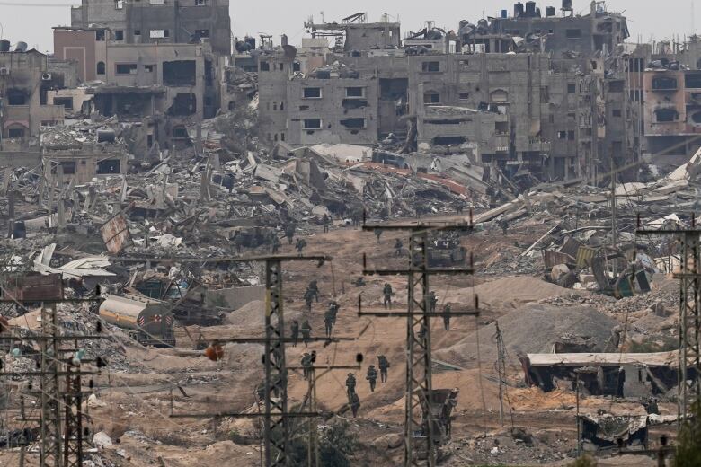 Soldiers are seen from a distance walking along a dirt path between destroyed buildings and power lines