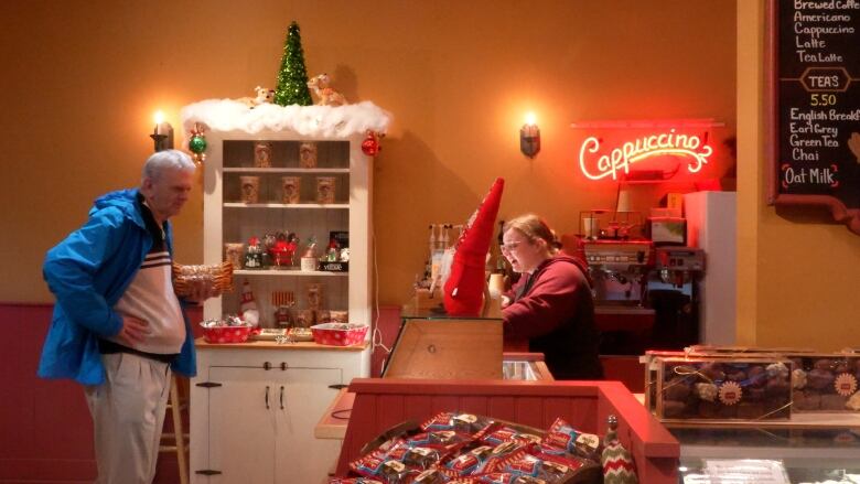 A man stands with his hands on his hips as he speaks with a person sitting at a cashier, with candies and cookies throughout the shop.