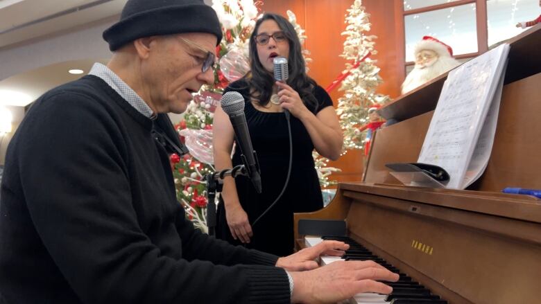 A man in his 60s is at the piano in a lobby adorned with Christmas decorations, playing alongside a female singer beside him as they perform for hotel guests in the Lobby of the Penticton Lakeside Resort.