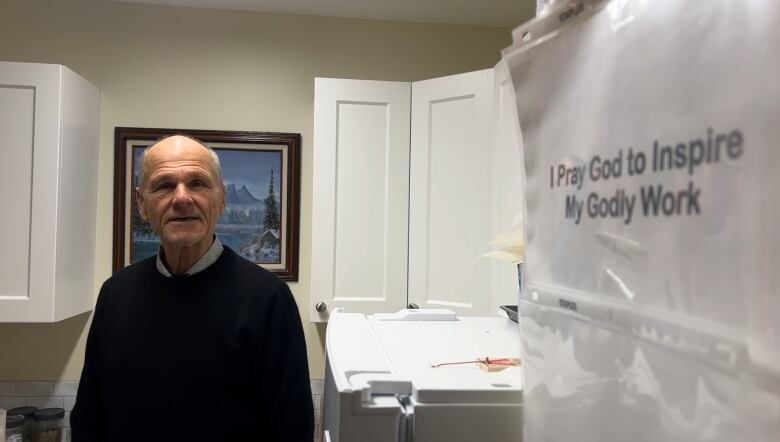 A man in his 60s smiles as he looks at the camera in the kitchen of a small apartment in Penticton, B.C. On the wall, and closer to the camera taking this photo, is a white sheet of paper with words printed on them reading, 