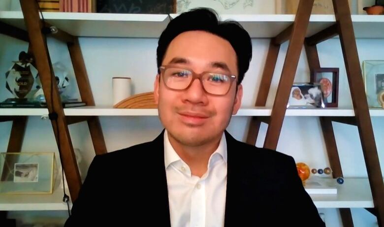 A man in glasses sits in front of a bookshelf.