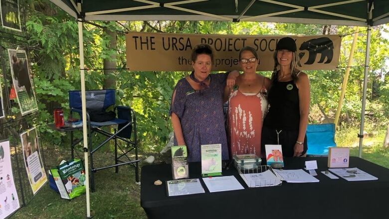 Three women stand behind a table with informational materials, with a sign 