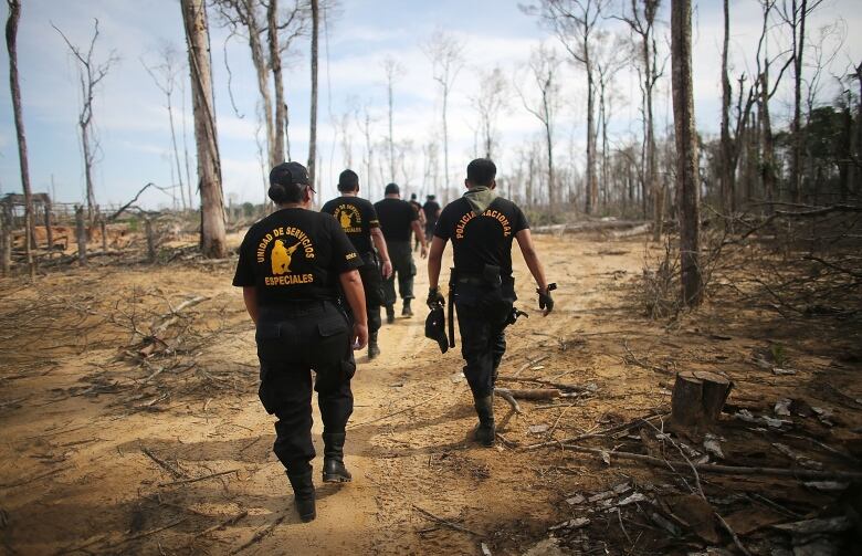 A group of men wearing black police uniforms with gold lettering march through a sandy area of the Amazon that's been stripped of trees and vegetation.