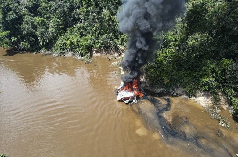 An aerial image shows a small structure engulfed in flames and smoke near the forested shore of a river flowing with brown water. 